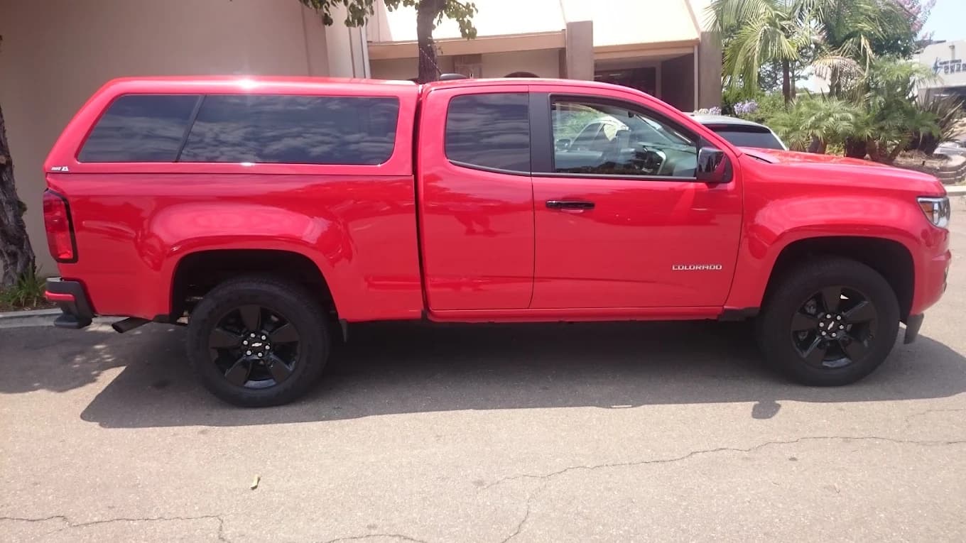 Red Chevrolet Colorado with a color-matched A.R.E. cap, parked beside the shop building