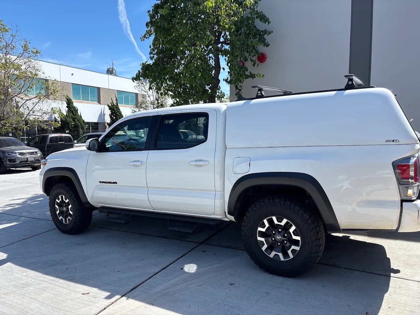 White Toyota Tacoma with an A.R.E. cap and roof crossbars, parked on the shop’s concrete apron